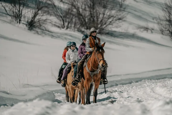 Group riding a horse in snowy landscape, led by person in cowboy hat.