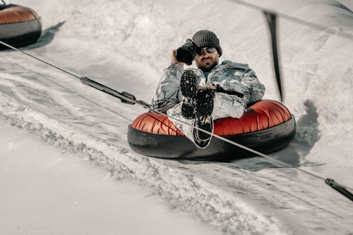 Person in winter clothes on an orange snow tube, holding a camera, being pulled up a snowy hill.