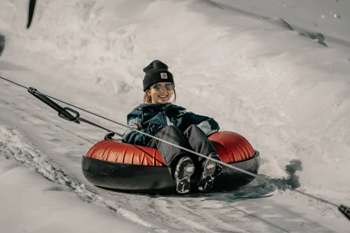 Person tubing down a snowy slope on a red tube wearing sunglasses and a black beanie.