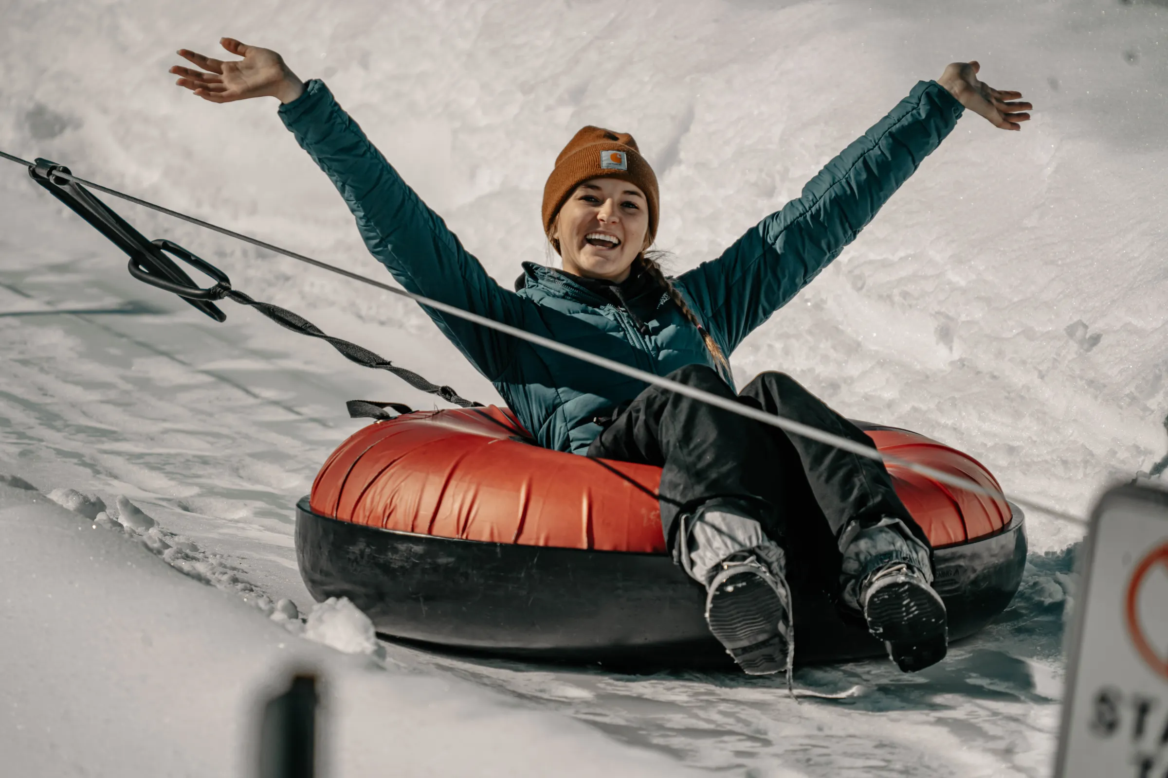 Person in winter clothing riding an inflatable tube on snow with raised arms, smiling.