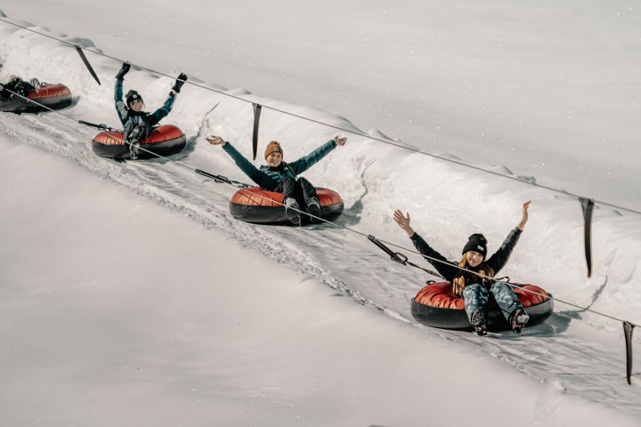 Three people on snow tubes holding ropes, sliding down a snowy slope with raised arms.