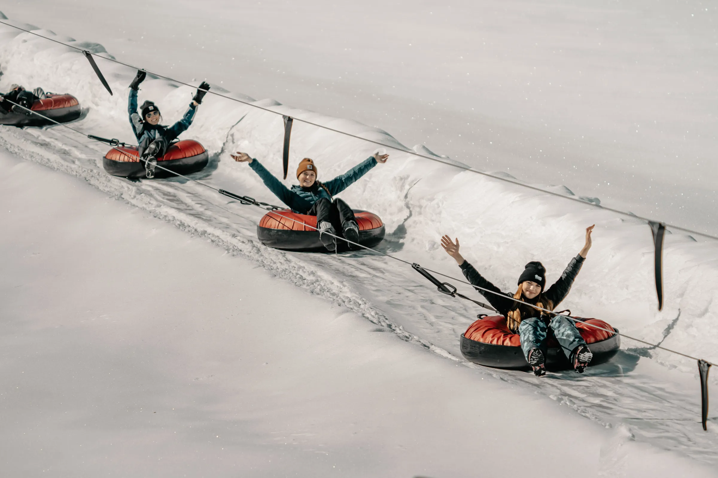 Three people on snow tubes holding ropes, sliding down a snowy slope with raised arms.