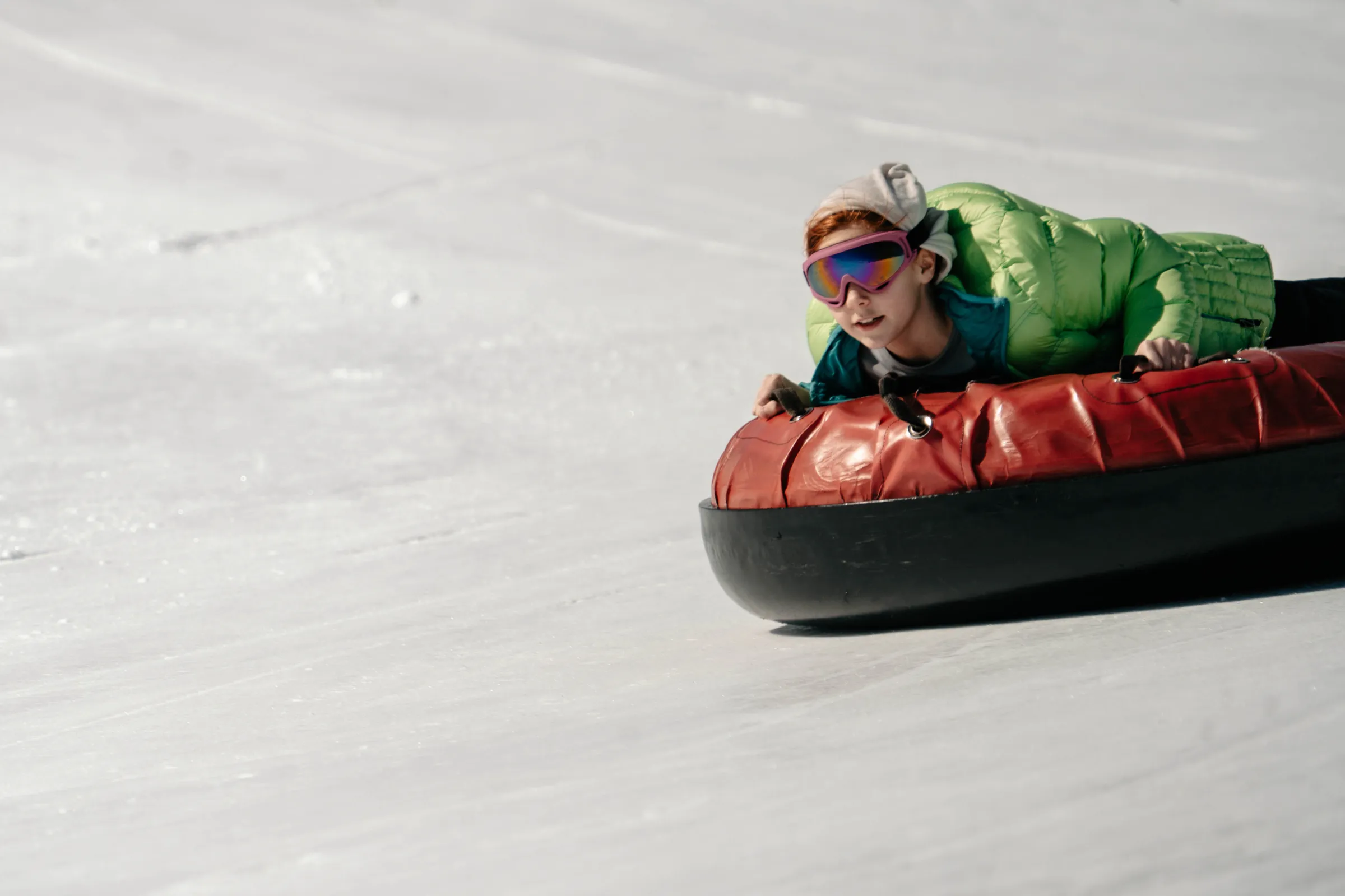 Child in green jacket sledding on a red tube down a snowy slope.
