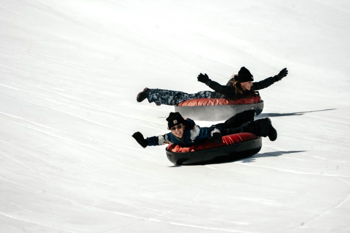 Two people snow tubing downhill on a snowy slope, arms outstretched.