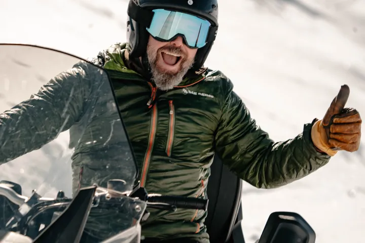 Man on snowmobile gives thumbs up, wearing helmet, goggles, and green jacket.