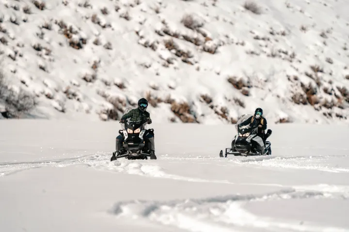 Two people riding snowmobiles on a snowy landscape with hills in the background.