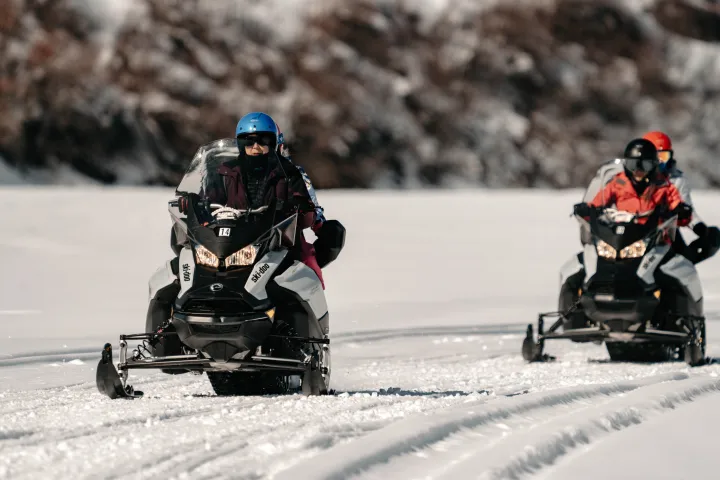 Two people riding snowmobiles on a snowy landscape with trees in the background.