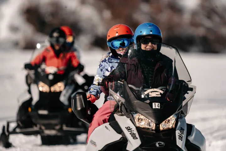 Three people riding snowmobiles on a snowy landscape, wearing helmets and winter gear.