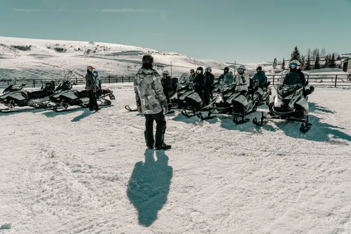 Group on snowmobiles in snowy landscape, listening to a person standing in front.