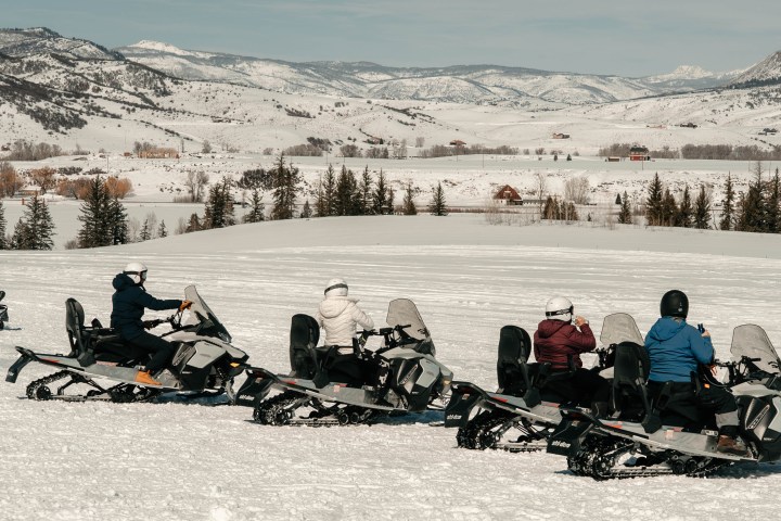 Snowmobilers riding in a snowy mountain landscape with trees and distant hills.