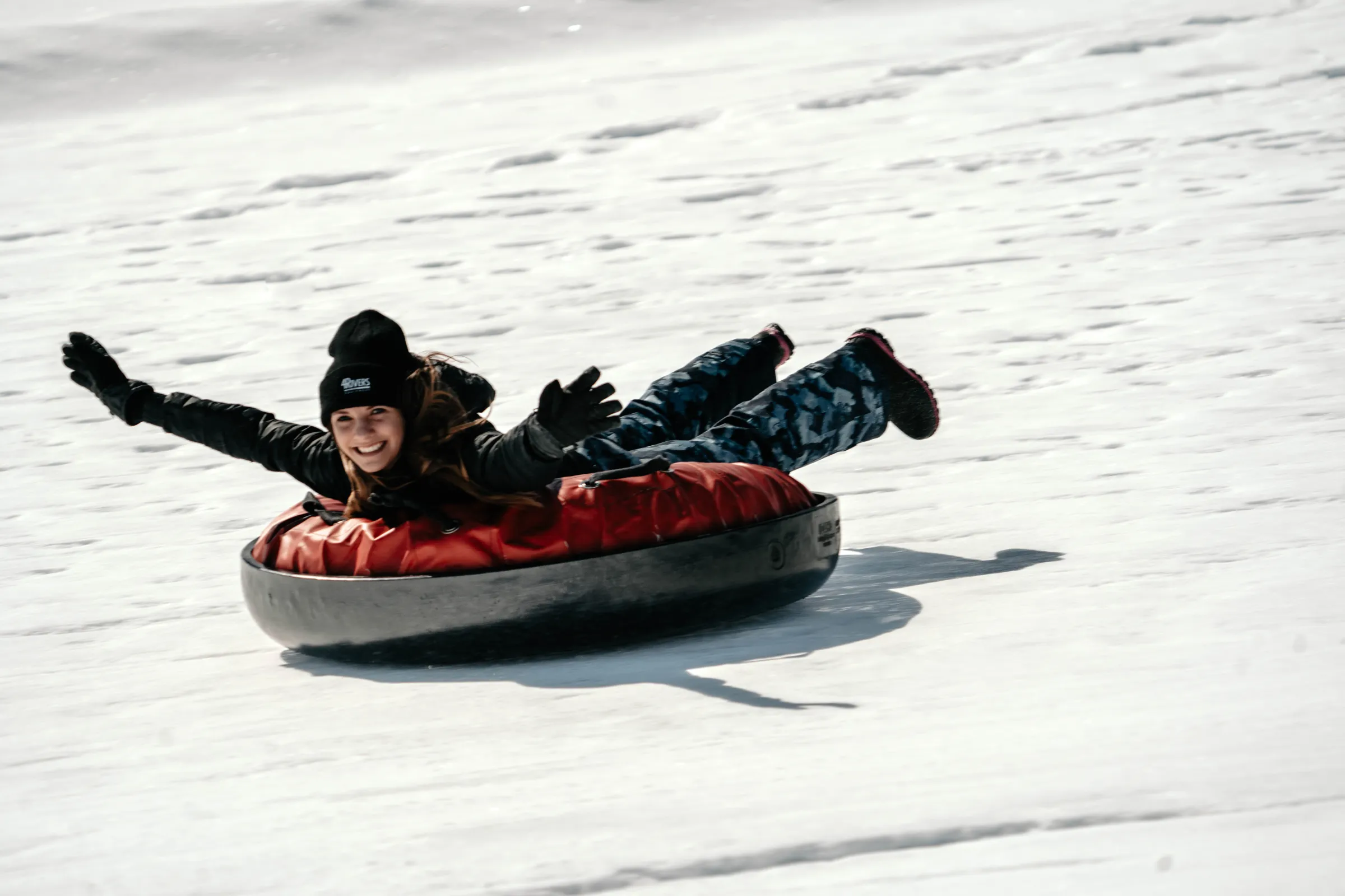 Person sliding on snow in a red tube with arms outstretched, smiling, wearing a black hat and gloves.