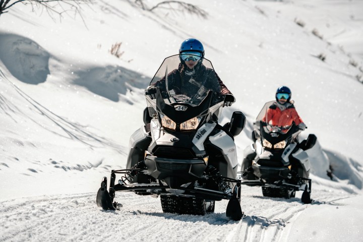 Two people riding snowmobiles on a snowy mountain trail, wearing helmets and winter gear.