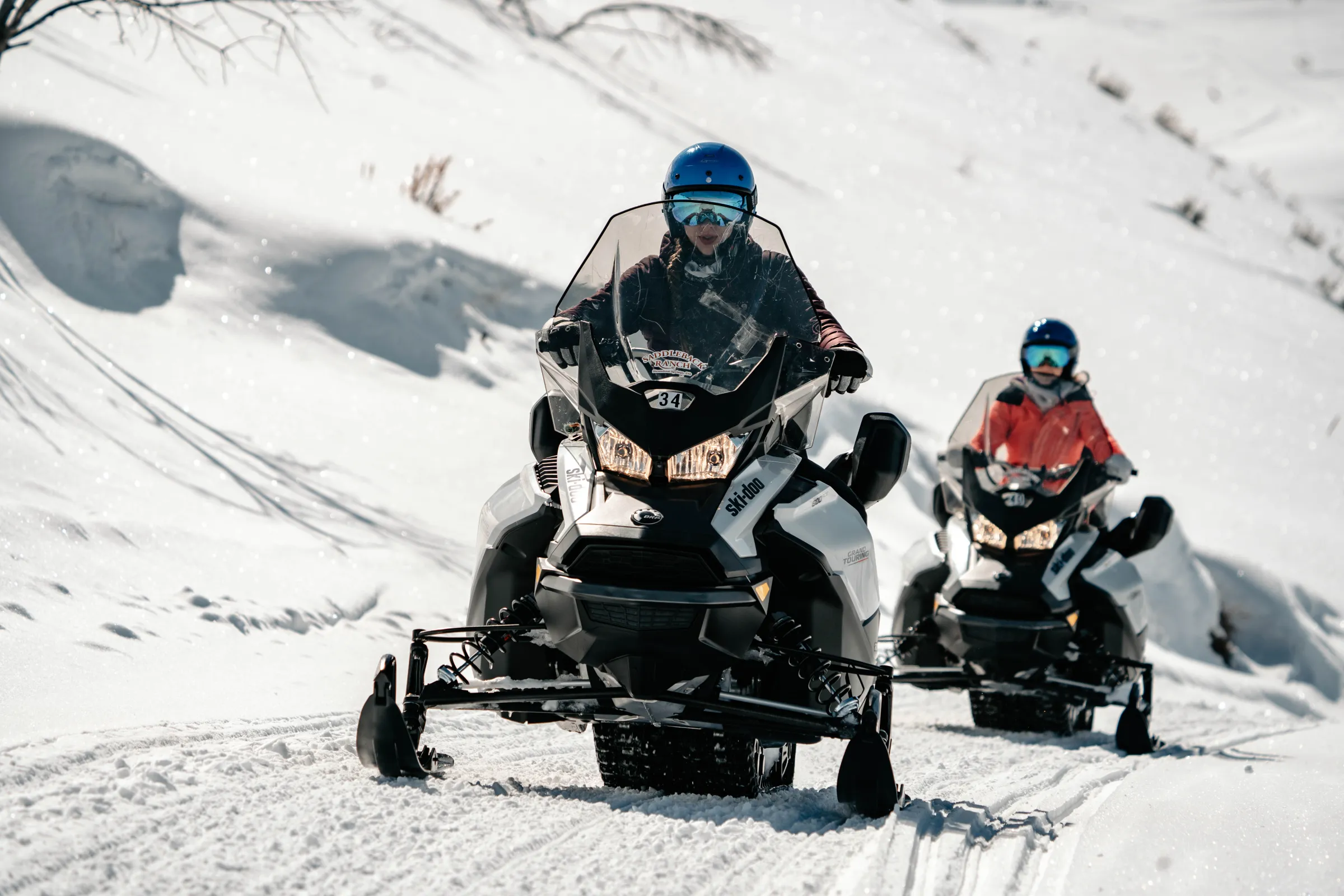 Two people riding snowmobiles on a snowy mountain trail, wearing helmets and winter gear.
