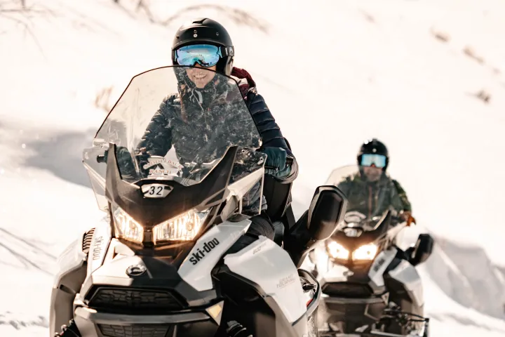 Two people riding snowmobiles on a snowy mountain path, wearing helmets and goggles.