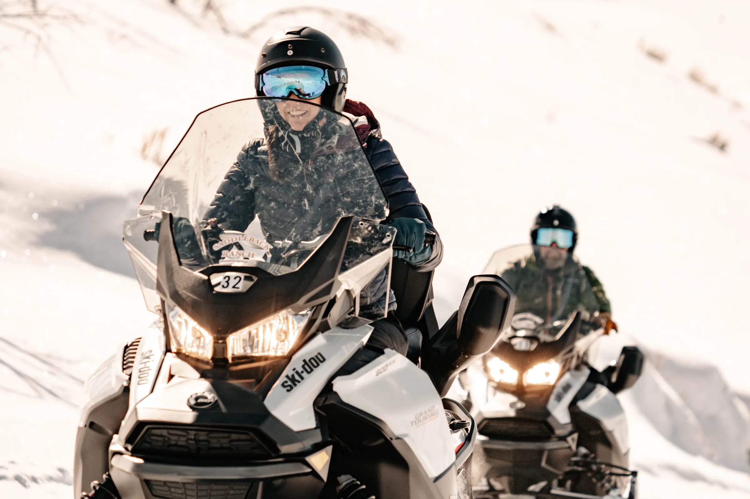 Two people riding snowmobiles on a snowy mountain path, wearing helmets and goggles.