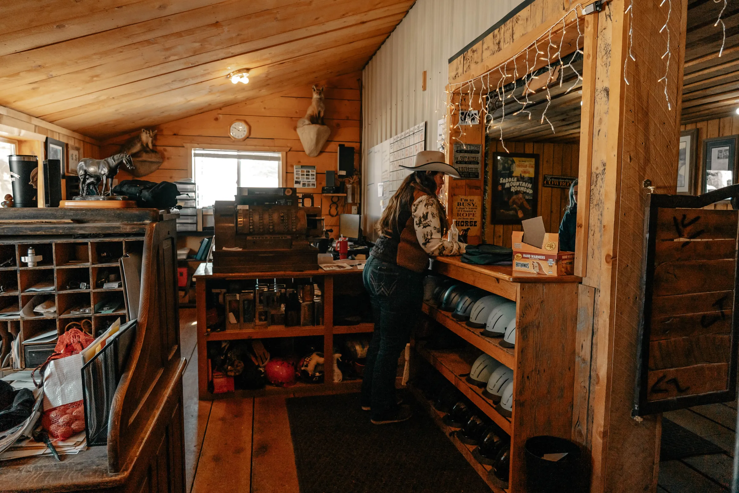 Person in cowboy hat at wooden counter in rustic store with helmets on shelves.