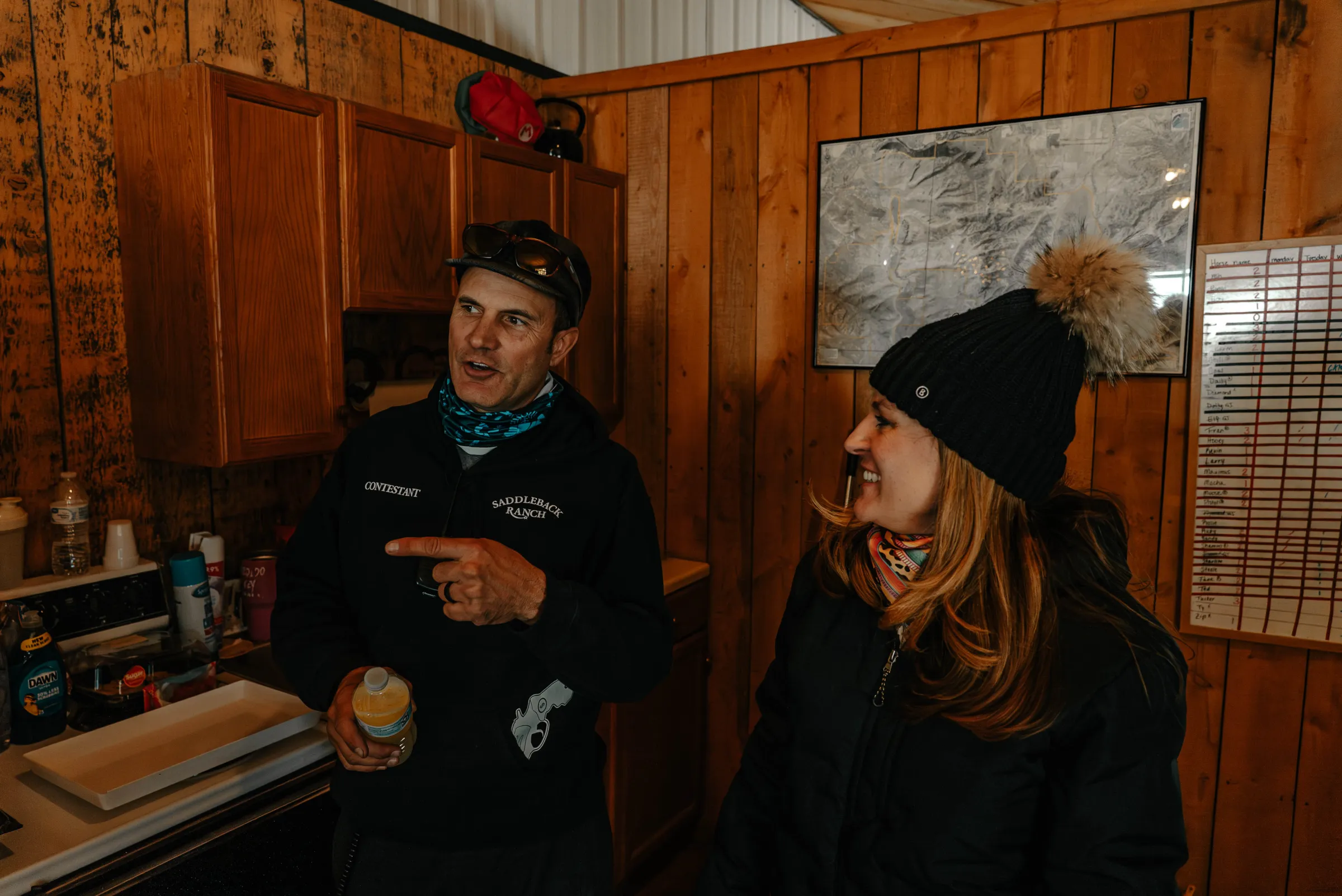 Two people in winter clothing talking inside a rustic wooden room.