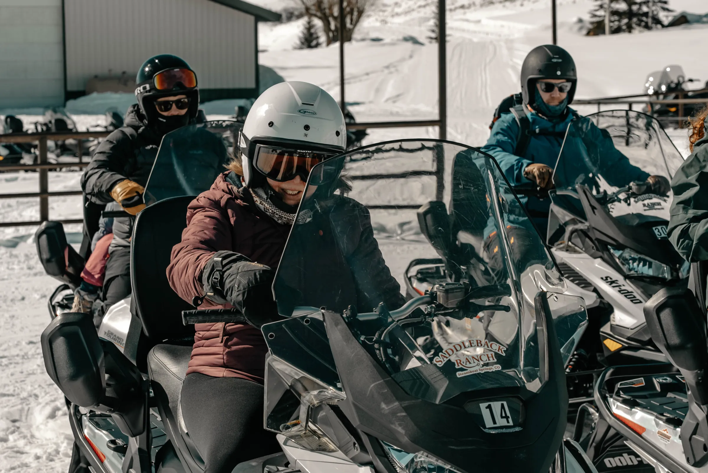 Three people in winter gear on snowmobiles in a snowy outdoor setting.
