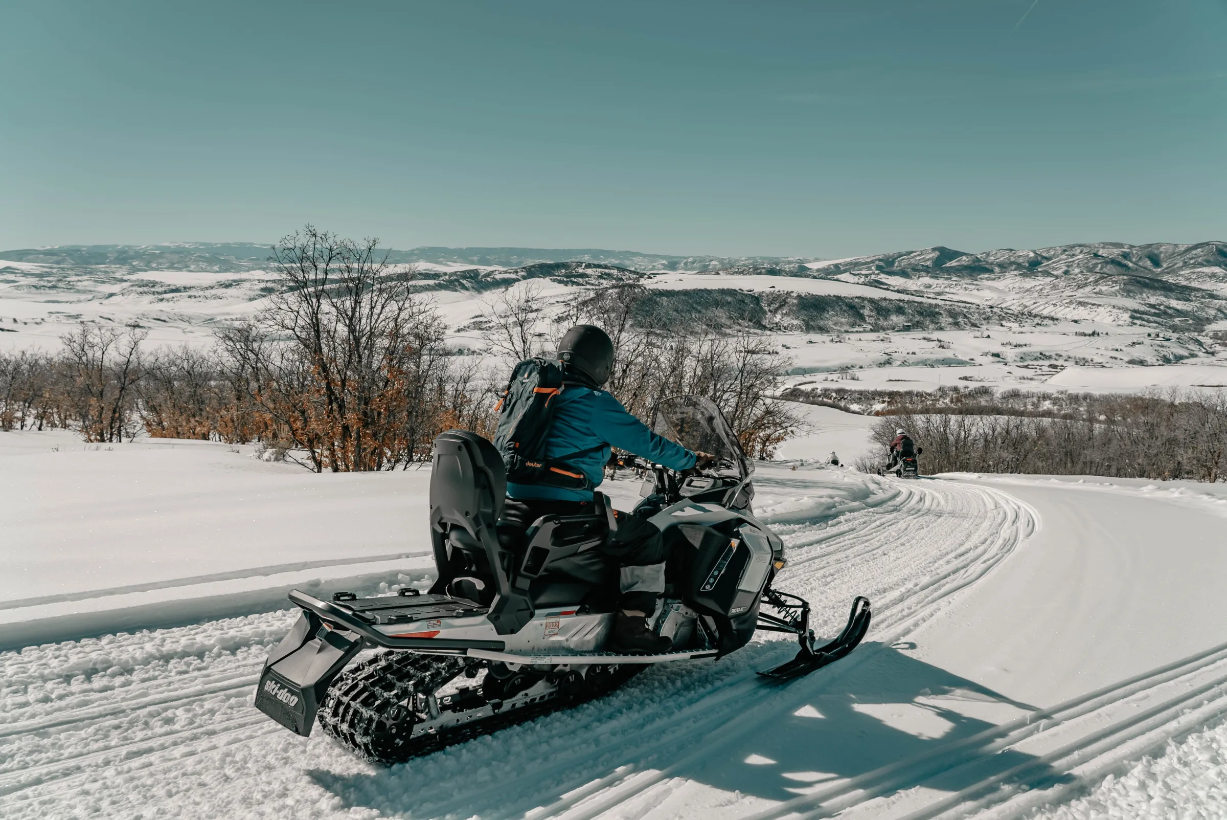 Person on a snowmobile driving on a snowy trail with mountains in the background.