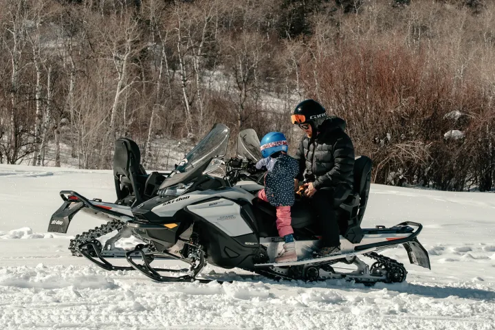 Adult and child wearing helmets on a snowmobile in snowy landscape.