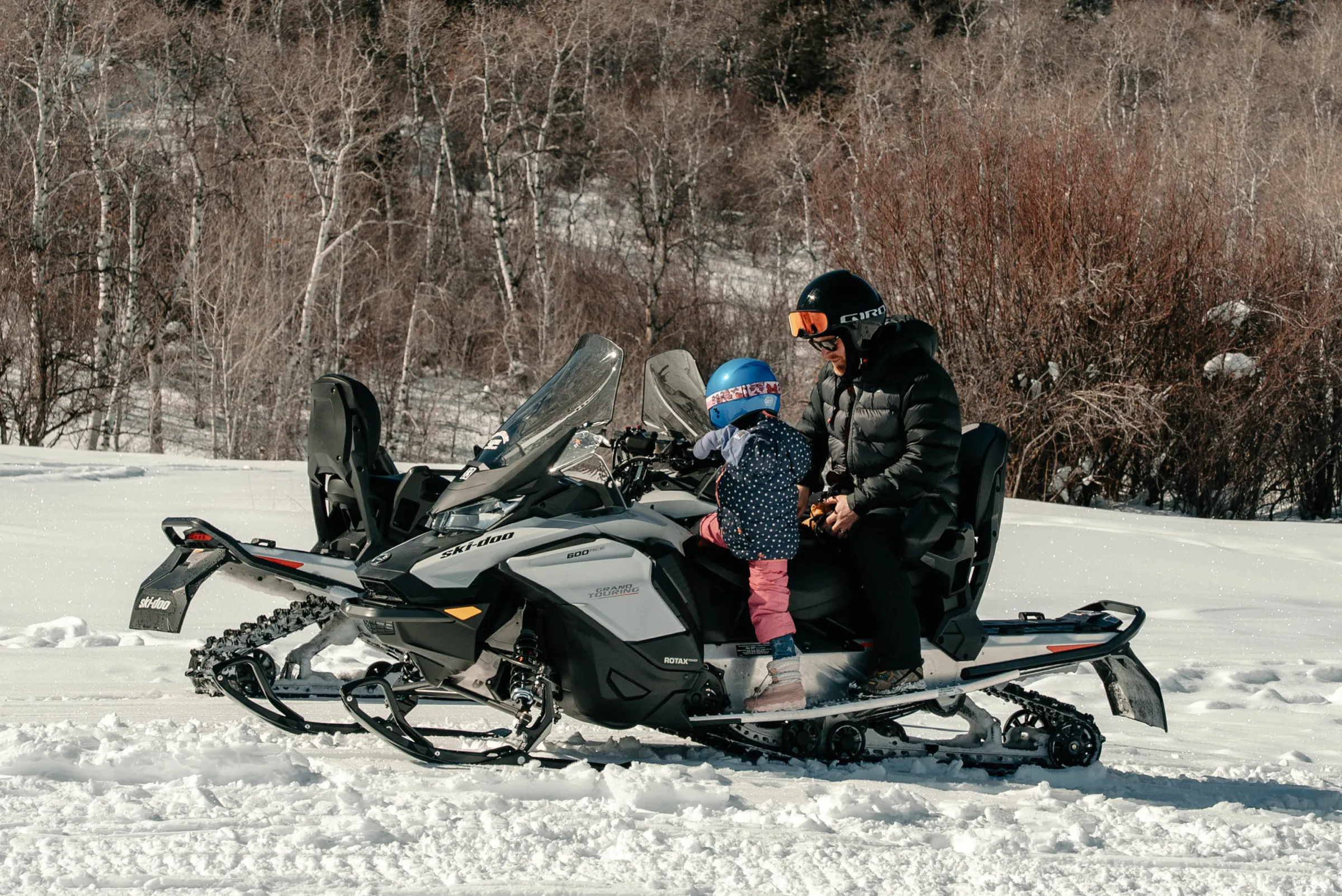 Adult and child wearing helmets on a snowmobile in snowy landscape.