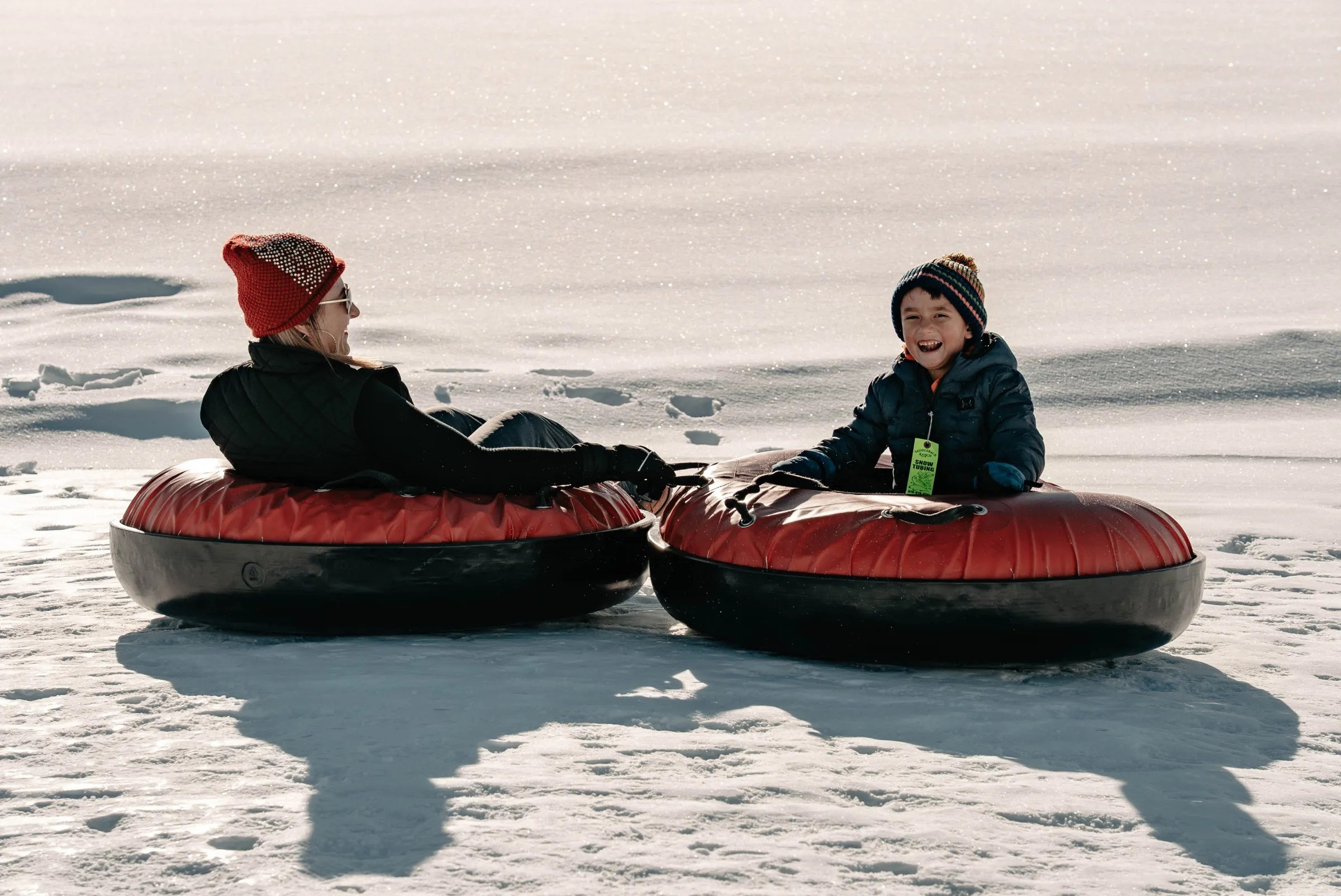 Two people smiling on red snow tubes in a snowy landscape.