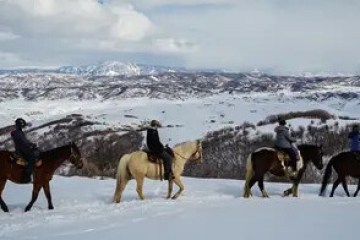 Four people horseback riding in snowy landscape with mountains in background.